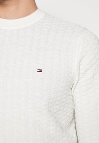 Pull en tricot blanc avec un motif de vagues, un col côtelé et un petit logo brodé en bleu marine et rouge sur la poitrine.