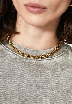 Collana in oro con maglie intrecciate, indossata sopra una maglietta grigia chiara con scollatura rotonda e texture morbida.