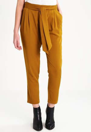 Kobieta w musztardowożółtych, wysokich, zwężanych spodniach z przodu wiązanych paskiem oraz czarnych botkach, stojąca na białym tle.