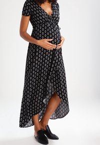 Robe de maternité noire avec un motif de diamants blancs, manches courtes, design enveloppant et ourlet asymétrique. Associée à des chaussures noires à enfiler.