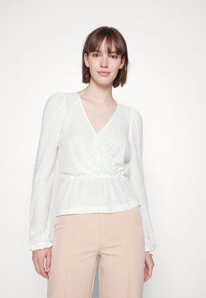 Blusa branca, texturizada, com mangas longas e design cruzado, apresentando uma cintura franzida. Combinada com calças bege claras.