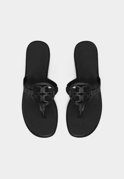 Ζευγάρι μαύρων δερμάτινων σανδάλων με λουράκια τύπου thong και στρογγυλή, περικομμένη λεπτομέρεια λογότυπου στα λουριά, εμφανιζόμενα σε λευκό φόντο.