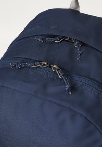 Niebieski plecak nylonowy z dwoma zasuwanymi kieszeniami z metalowymi elementami i ozdobnymi sznurkami. Teksturowana powierzchnia zapewniająca trwałość.