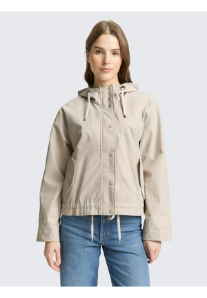 Junge Frau trägt eine beige Kapuzenjacke mit Kordelzug und blaue Jeans, steht vor einem schlichten hellen Hintergrund.