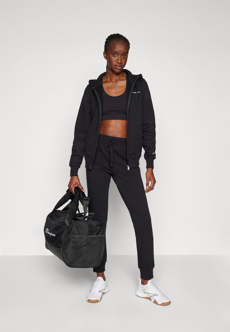 Felpa nera con zip e cordino, abbinata a pantaloni joggers neri e una borsa duffel nera. Scarpe da ginnastica bianche completano il look.