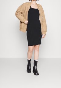 Abito nero a costine abbinato a una giacca bomber color sabbia. La modella indossa stivaletti neri alla caviglia con dettagli bianchi, mostrando un look elegante e moderno.
