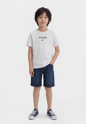 Garçon debout portant un t-shirt blanc Nike "PARIS", un short en jean bleu et des baskets bleu marine sur un fond blanc uni.