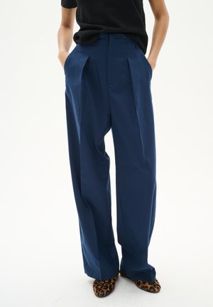 Wide-leg navy trousers made of a smooth fabric, featuring front pleats and side pockets, paired with animal print shoes.