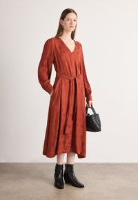Rostfärgad klänning med långa ärmar och V-ringning, knytbälte i midjan och subtil präglad mönster. Bärs med svarta ankelstövlar och en texturerad handväska.