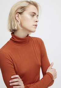 Rostfarbener Rollkragenpullover aus weichem Stoff, taillierte Form mit langen Ärmeln. Silberne Creolen setzen einen schlichten Akzent.