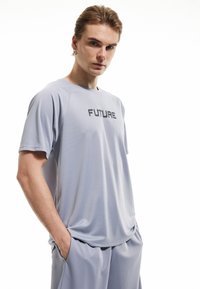 Camisa deportiva de color gris claro con la palabra "FUTURE" impresa en negro. Presenta mangas cortas y un corte holgado, confeccionada con tela transpirable.