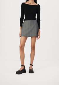 Vrouw in een zwarte off-shoulder top, grijze gestreepte mini-skirt en zwarte enkelband chunky platform schoenen die op een witte vloer staat.