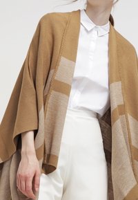 Bruin en beige oversized vest met een textuurontwerp, voorzien van een brede open voorkant en losse mouwen, gecombineerd met een wit overhemd.