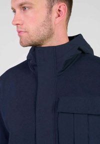 Sudadera azul marino con cuello alto, tejido texturizado y un bolsillo frontal. Presenta un diseño minimalista con cierres ocultos.