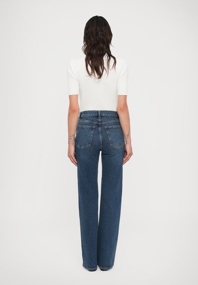 Rifle s vysokým pásom a širokými nohavicami v tmavomodrom denime s piatimi vreckami, jemným prešívaním a hladkou textúrou. Pohľad zozadu ukazuje výraznú siluetu.