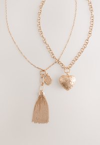 Two gold necklaces, one with a heart-shaped engraved pendant and thick chain, the other with a tassel and small charm on a fine chain.