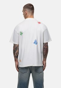 Witte katoenen t-shirt met multikleurige vlinderafdrukken in groen, blauw, rood en oranje op de rug. Ronde hals en korte mouwen.