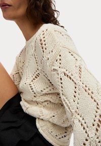 Off-white gebreide trui met een diamantpatroon, open stitch ontwerp en brede mouwen. Gepaard met een zwarte gerimpelde kledingstuk.