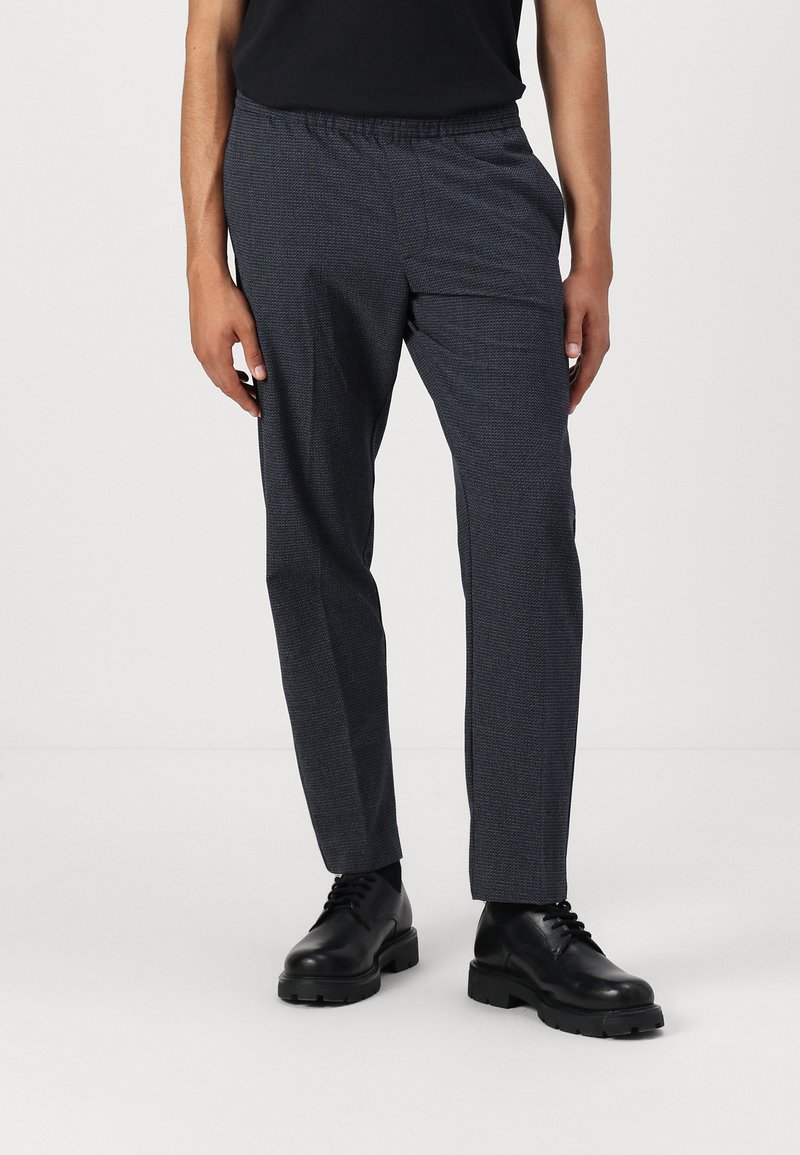 Pantalones oscuros de rayas grises para hombre con un ajuste relajado, que cuentan con una cintura elástica y una textura sutil, usados con zapatos negros de cordones.
