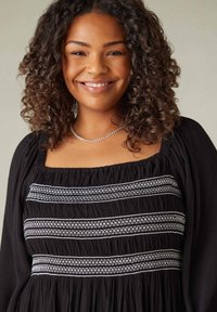 Svart klänning med smockad byst och vita broderade mönster, fyrkantig halsringning och lång ärmar. Lockigt hår och enkel silverhalsband.