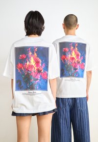 Hvite bomull T-skjorter med et stort blomstermotiv på baksiden, med rosa roser og blå flammer, rammet inn av en rektangulær kant.