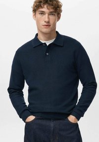 Granatowy, długi rękaw, koszulka polo z fakturowanym splotem, trzy guziki i klasyczny kołnierzyk. Noszona casualowo, z rękami w kieszeniach.