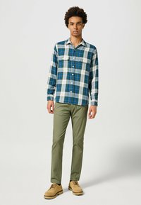 Camisa de franela para hombre en cuadros azules y verdes, con mangas largas, dos bolsillos en el pecho y cierre frontal de botones. Combinada con pantalones verdes y zapatos color beige.