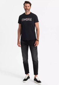 Sort t-shirt med "OMBRE" tekstdesign i multifarver, parret med mørkegrå slim-fit jeans og sorte sneakers med hvide såler.