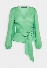Blouse portefeuille verte à manches longues, en tissu texturé, taille peplum et détail noué. Le décolleté en V met en valeur la silhouette.