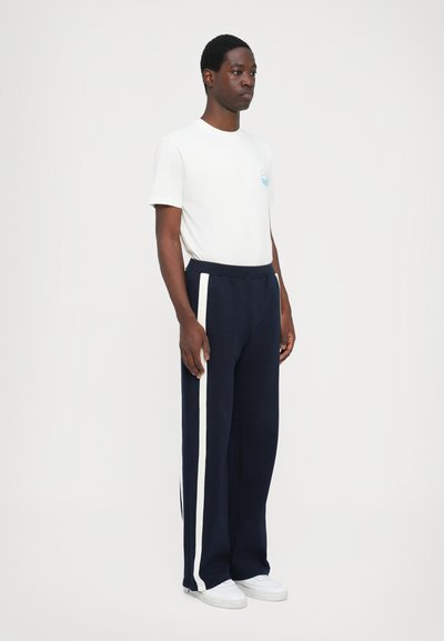 White short-sleeve t-shirt with a graphic on the chest, paired with navy trousers featuring white side stripes and a relaxed fit.