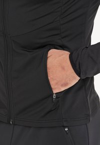 Sort zip-up jakke med glat, strækbart stof, flade sømme, sidelommer med lynlåse og en tætsiddende design.