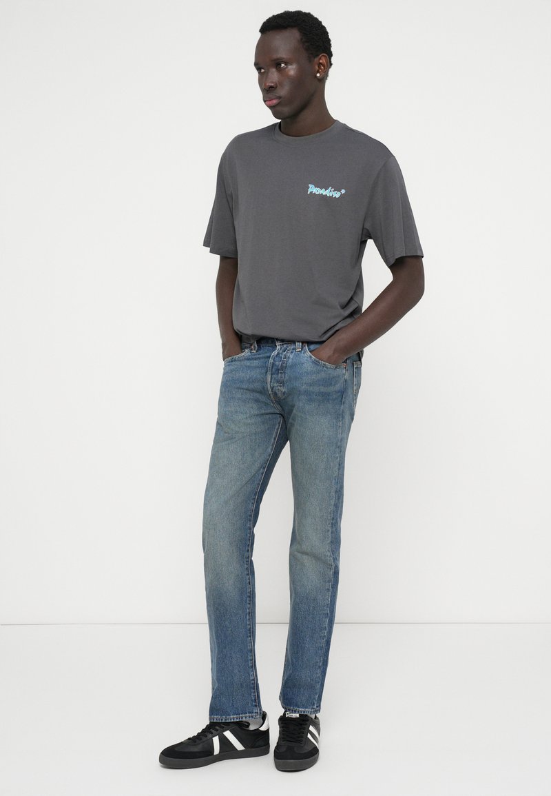 Camiseta de algodón gris con mangas cortas que presenta un logo azul, combinada con unos vaqueros de denim azul claro y zapatillas negras con rayas blancas.