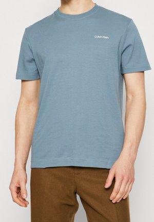 Mand iført en dæmpet blå Calvin Klein t-shirt med korte ærmer og brune bukser, stående mod en lys baggrund.