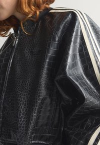 Crna jakna od umjetne kože s teksturiranim uzorkom krokodilske kože, s bijelim bočnim prugama i zatvaračem. Glatka završna obrada i podstavljeni dizajn.