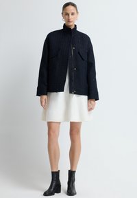 Granatowa kurtka o fakturze z złotymi guzikami i dwoma przednimi kieszeniami, noszona na białej spódnicy, w połączeniu z czarnymi botkami za kostkę.