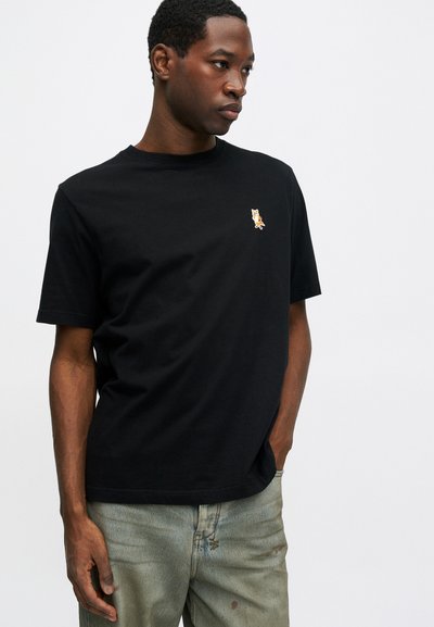 Joven vestido con una camiseta negra con un pequeño logo de un zorro en el lado izquierdo del pecho, combinada con unos jeans gris claro, de pie frente a un fondo blanco.