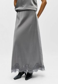 Longue jupe grise en satin avec un ourlet en dentelle détaillée. Le tissu a une texture lisse et s'écoule doucement. Associée à des chaussures noires à bouts pointus.