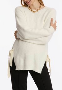 Weißer Strickpullover mit seitlichen Bindebändern. Weiche Textur, lockere Passform, gerippter Stoff und lange Ärmel. Kombiniert mit schwarzen Hosen.