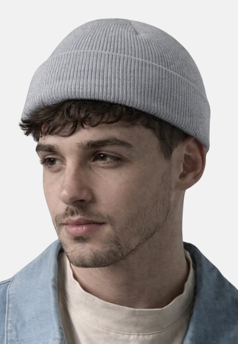 Junger Mann mit kurzen lockigen Haaren, der eine hellgraue gerippte Mütze, ein beigefarbenes Hemd und eine hellblaue Jacke trägt und leicht zur Seite blickt.