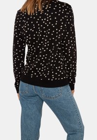 Maglione nero con motivo floreale bianco, polsini e orlo a costine. Abbinato a jeans in denim blu, che mostrano una vestibilità rilassata e una texture chiara.