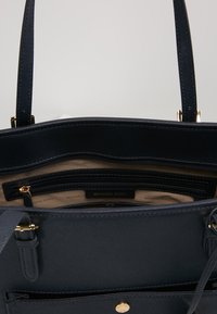 Sac à main noir Michael Kors avec ouverture supérieure montrant la doublure intérieure beige, une poche intérieure zippée et des détails en métal doré.