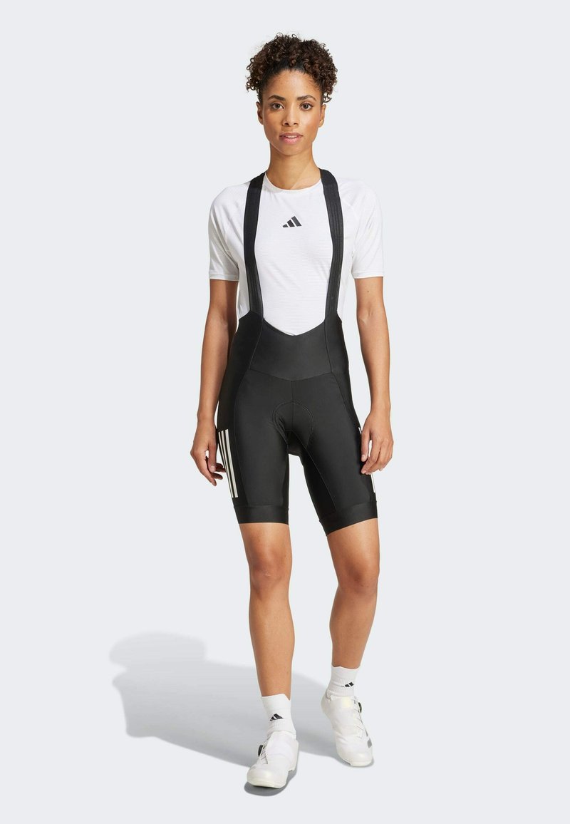 Schwarze Radfahr-Bib-Shorts mit gepolstertem Sitz, verstellbaren schwarzen Trägern und drei weißen Adidas-Streifen auf der linken Seite. Weißes T-Shirt mit kurzen Ärmeln.