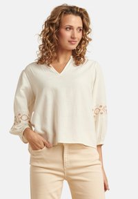 Cremfarbene Bluse mit V-Ausschnitt, weitem Ärmeln und bestickten Akzenten an den Bündchen. Der Stoff wirkt weich und leicht.