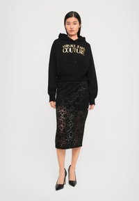 Svart avkortet hettegenser med "Versace Jeans Couture" i gullbokstaver, kombinert med et svart blond midi-skjørt og svarte spisse hæler.