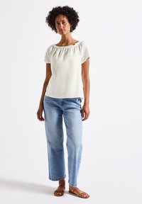 Top in lino avorio a maniche corte con scollatura arricciata, abbinato a jeans a gamba larga di un blu chiaro con dettagli a bottoni laterali e sandali flat.