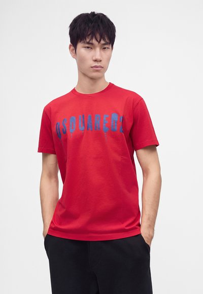 Jeune homme portant un t-shirt rouge avec un texte bleu "DSQUARED2", mains dans les poches, debout devant un fond clair uni.