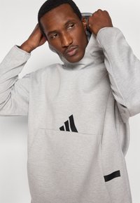 Pilkas džemperis su gobtuvu, didelė kangaroo tipo kišenė ir juodas trijų dryžių logotipas priekyje. Pagamintas iš minkšto audinio su laisvu siluetu.