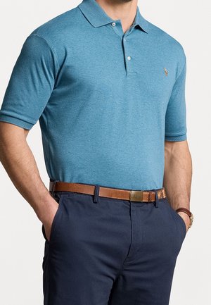 Homme portant un polo à manches courtes bleu, un pantalon marine et une ceinture en cuir marron, les mains dans les poches.