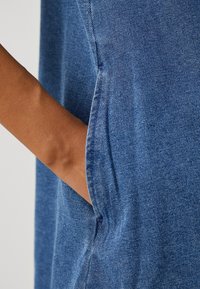 Denimvaate, jossa on syvän sininen sävy, näkyvä teksturoitu kudos ja lähikuva sivutaskusta, johon osa käsivarresta on työntynyt.