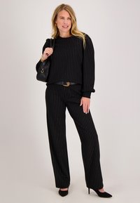Zwart pinstripe broekpak met top met lange mouwen en wijd uitlopende broek. Heeft een getailleerde taille met een riem en zwarte hoge hakken.